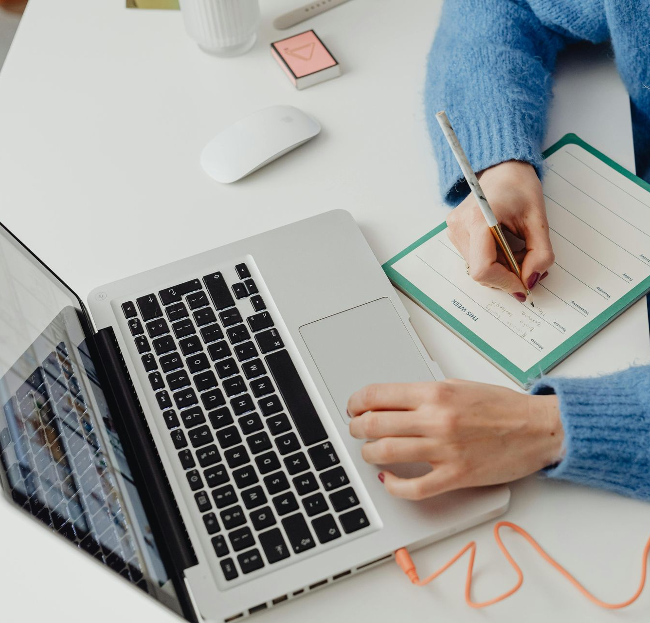 Person writing in notebook at a desk, next to a laptop, mouse, and other supplies.