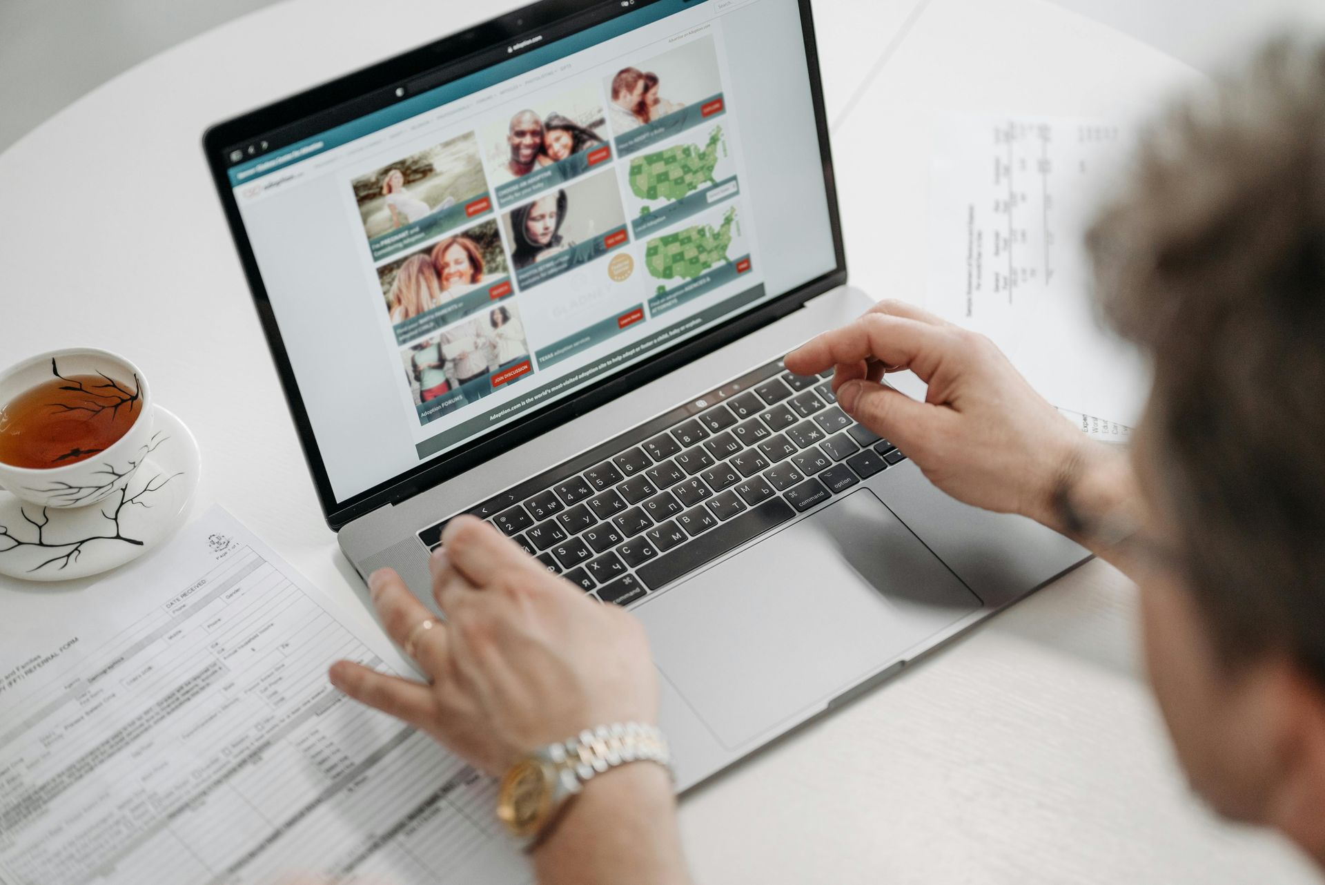 Person using a laptop, browsing a website with photos, tea and documents on the table.