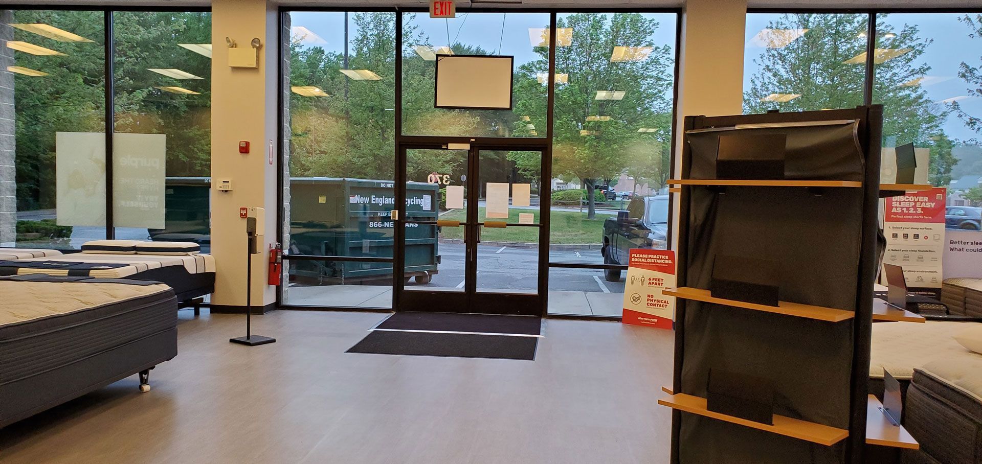Interior view of a store with large windows, double doors, and shelving units. Displays a bed, and the outside is visible.