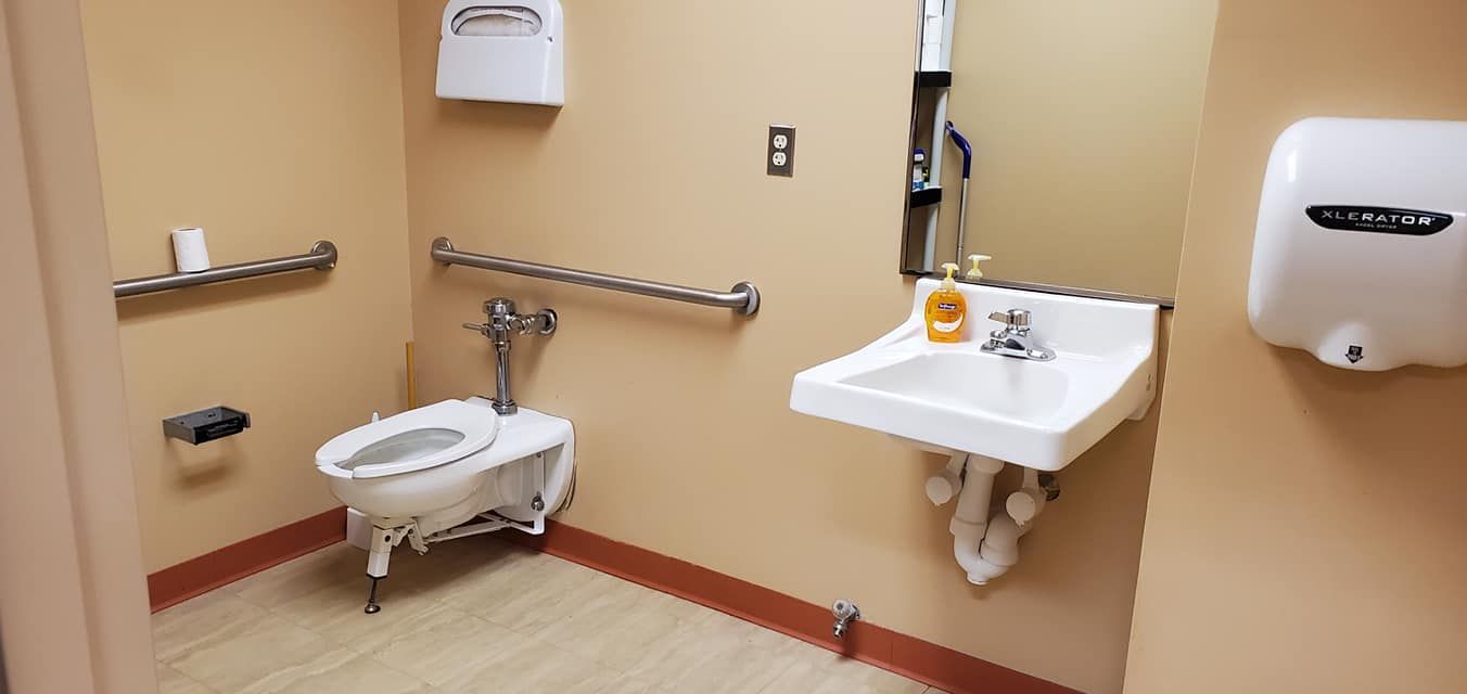 Accessible bathroom with toilet, sink, grab bars, and hand dryer. Neutral tan walls and flooring.
