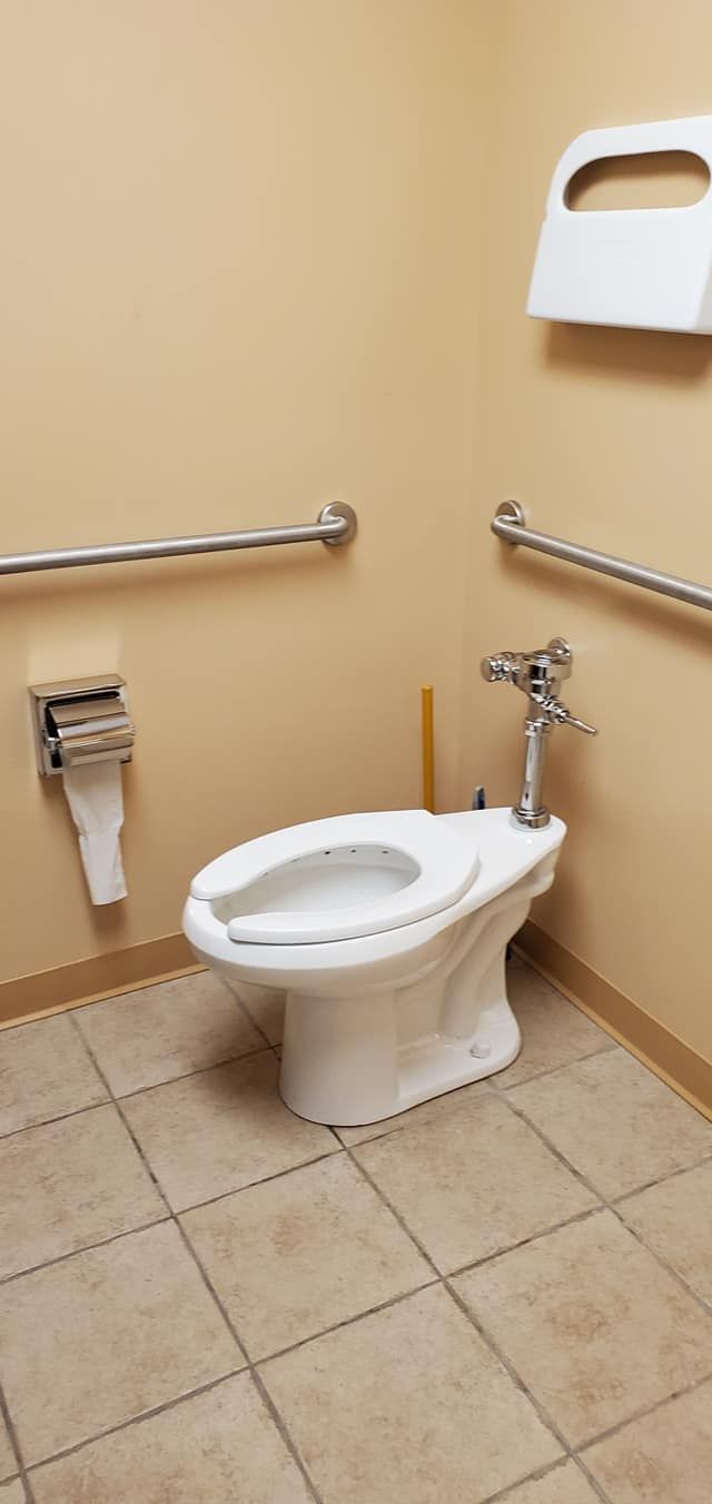 Public restroom with toilet, grab bars, and paper towel dispenser. Tile floor, beige walls.
