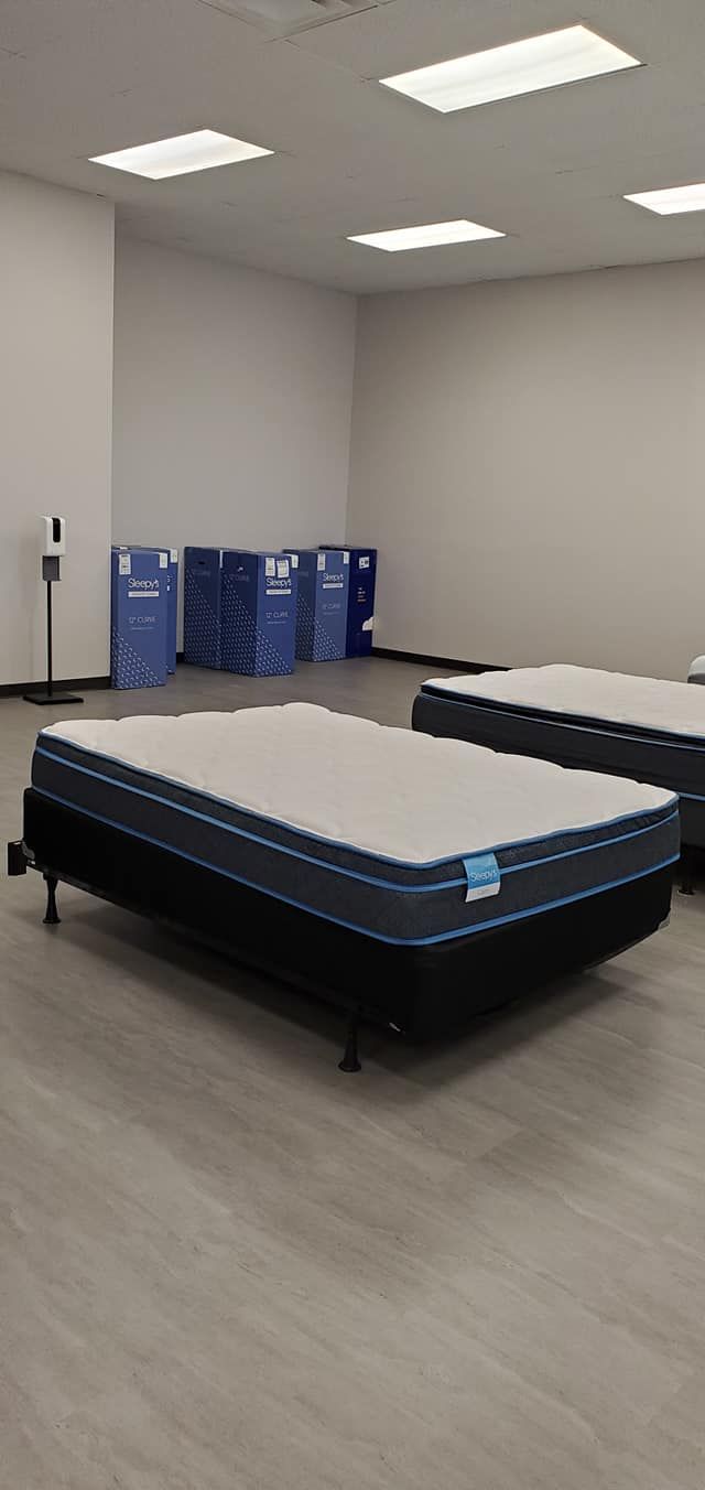 A mattress showroom with beds on display and boxes in the background. White walls, wood floor, bright overhead lighting.