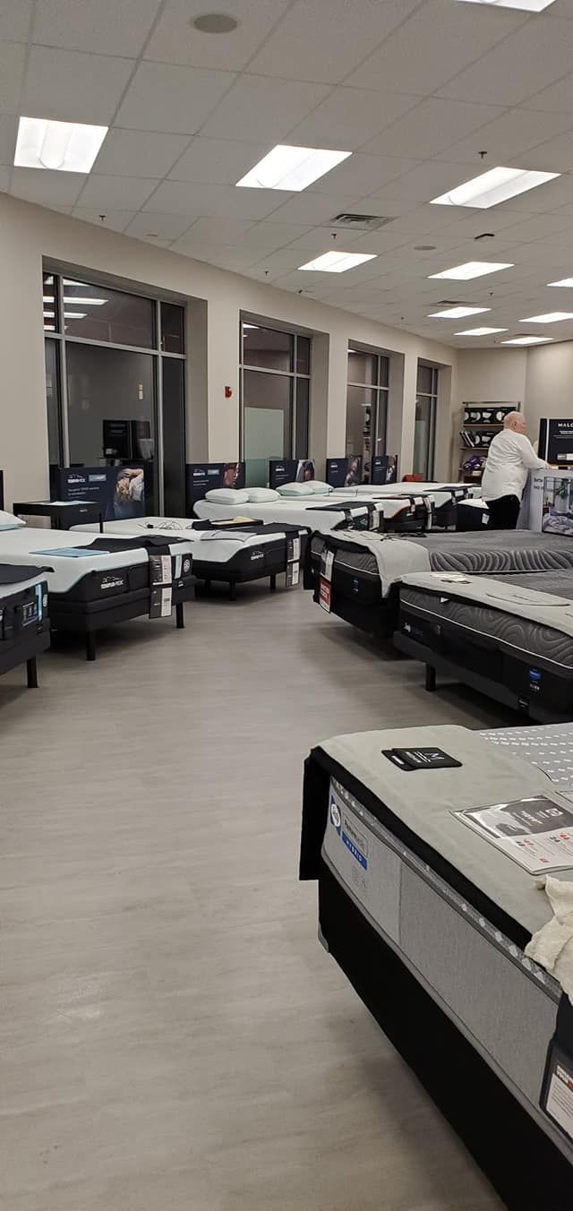 Mattress store interior with rows of beds on display, a person standing near the back, and large windows.