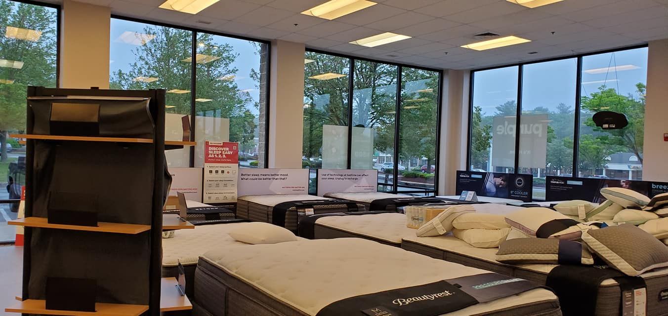 Mattress store interior with several beds displayed, large windows with a view of greenery.