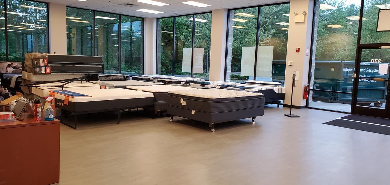 Interior of a mattress store with many mattresses on display. Large windows reveal a view of trees outside.