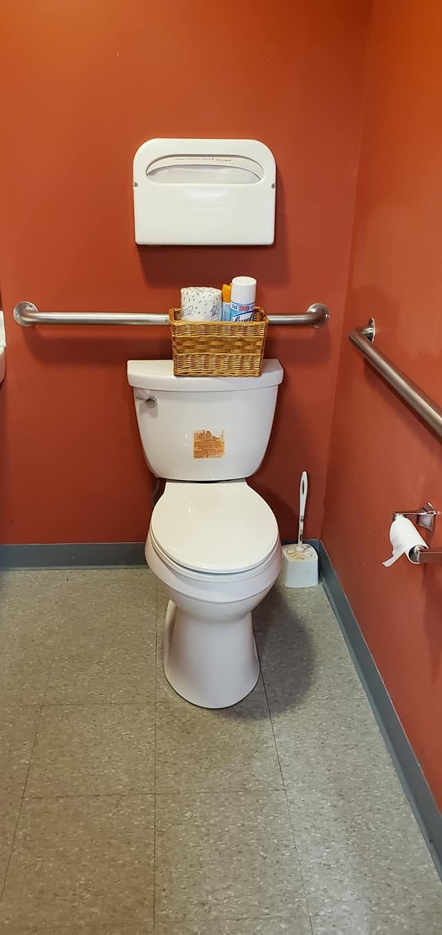 A toilet in a public restroom with an orange wall, handrails, and a basket of supplies on the tank.