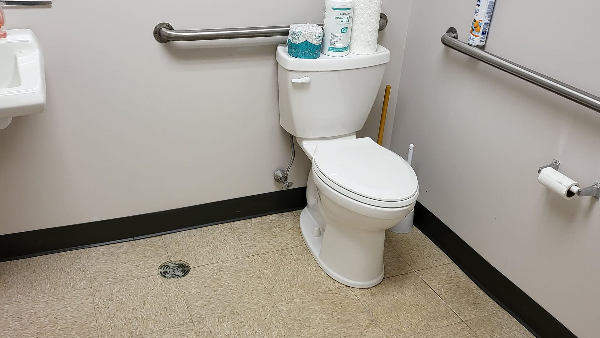 White toilet in a public restroom with grab bars, toilet paper holder, and floor drain.