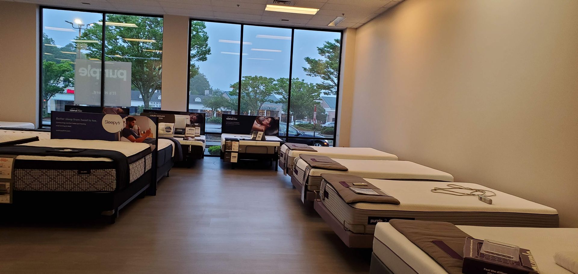 Mattress store interior with multiple beds displayed, large windows with outdoor view, and a light-colored wall.