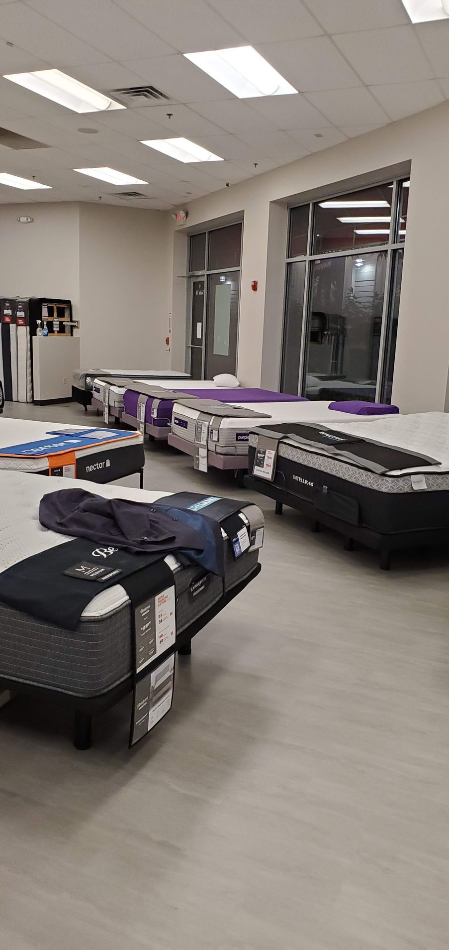 Mattress store interior with various mattresses on display, gray flooring, and bright overhead lights.