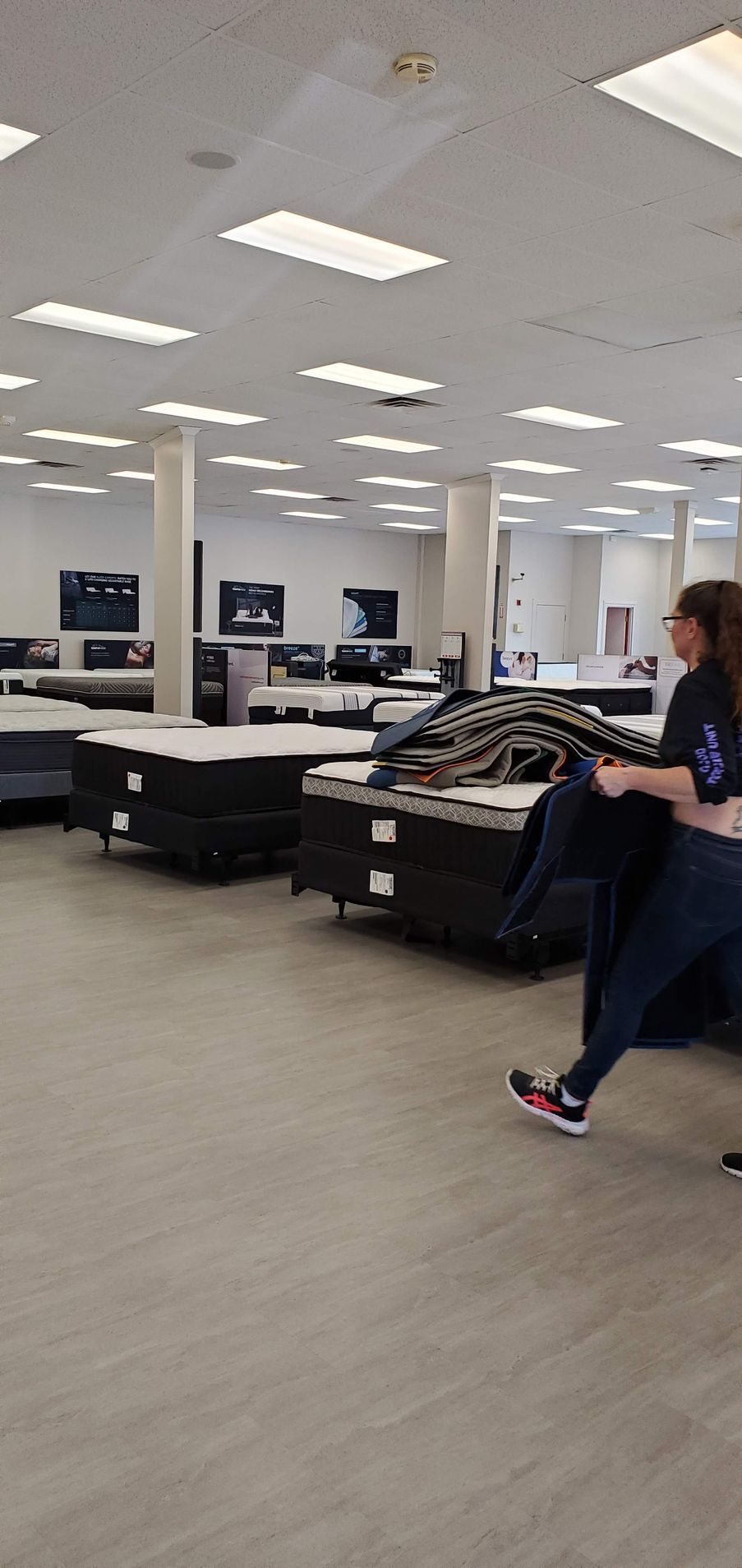 A person walks through a mattress store, holding a jacket, past rows of mattresses.