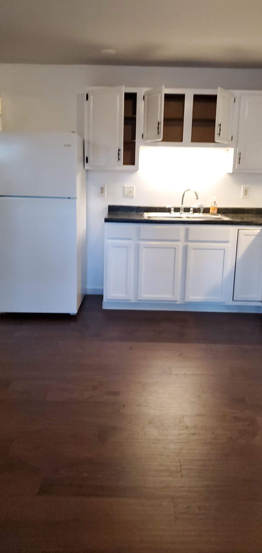 A small kitchen with white cabinets, a black countertop, and a white refrigerator on a dark wood floor.
