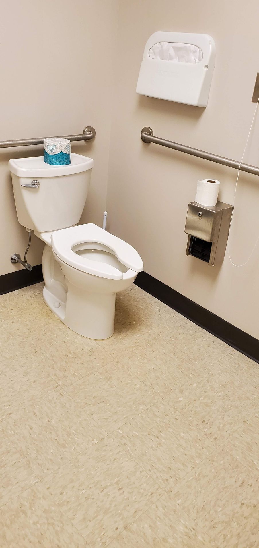 A white toilet in a public restroom with grab bars, a paper towel dispenser, and a roll of toilet paper.