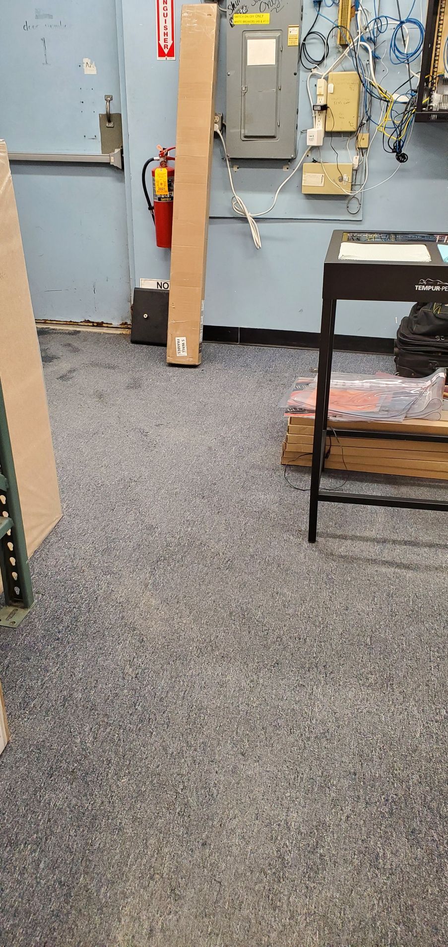 A warehouse interior with blue speckled flooring, a tall cardboard box, fire extinguisher, and electrical panels.