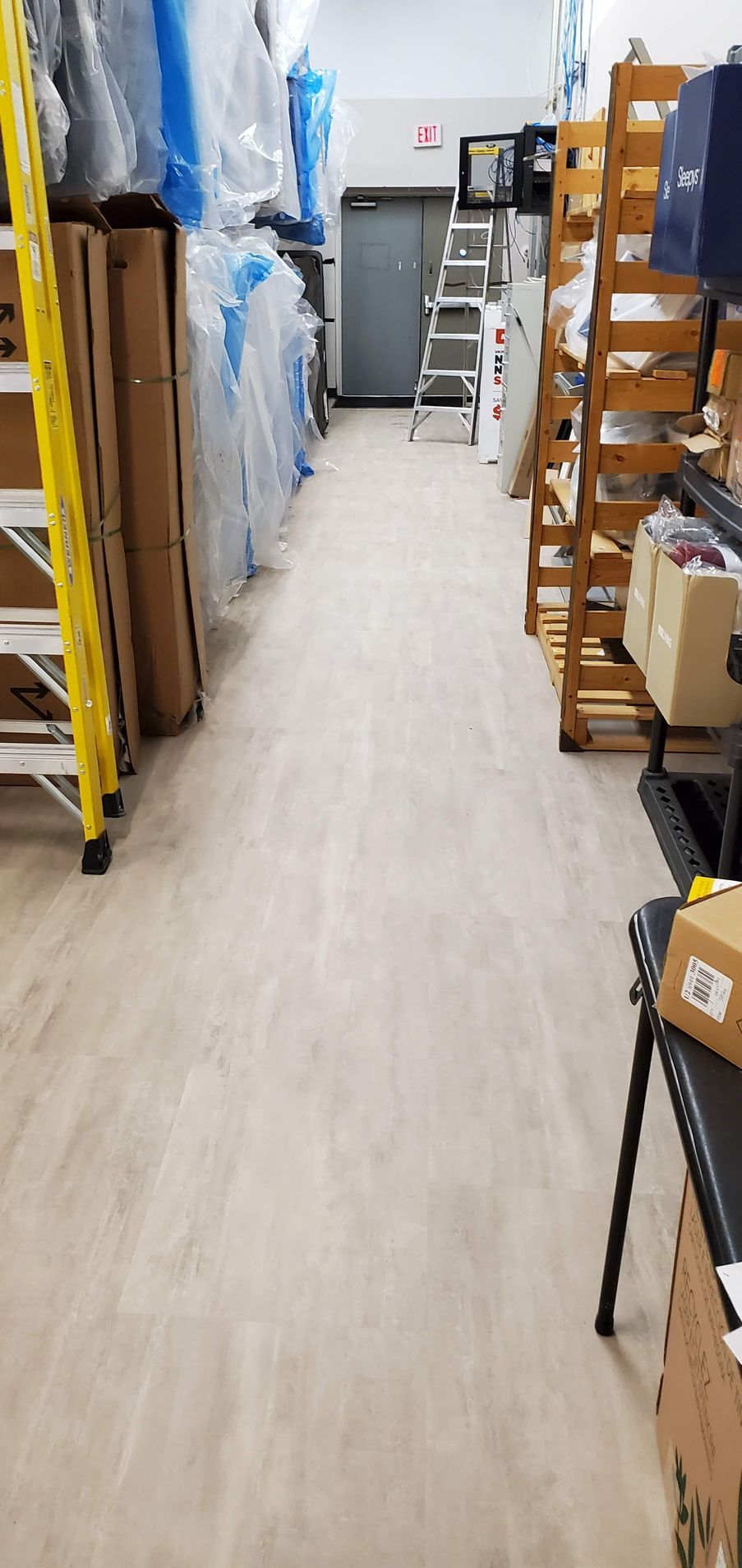 A narrow warehouse aisle with shelves, boxes, and a ladder leading to a door.