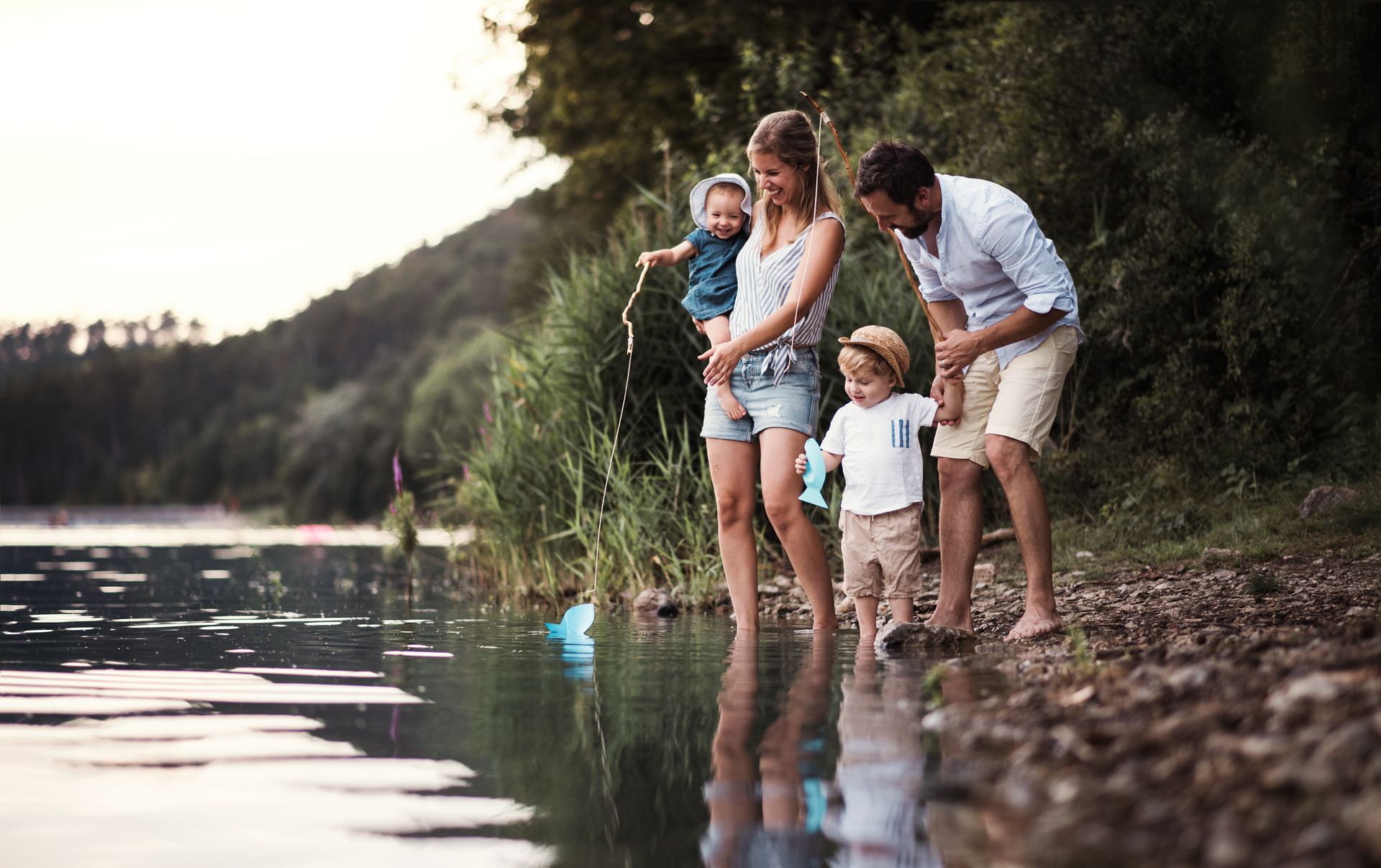 A family stands at the water's edge of a lake at sunset, watching a small blue toy boat float nearby.