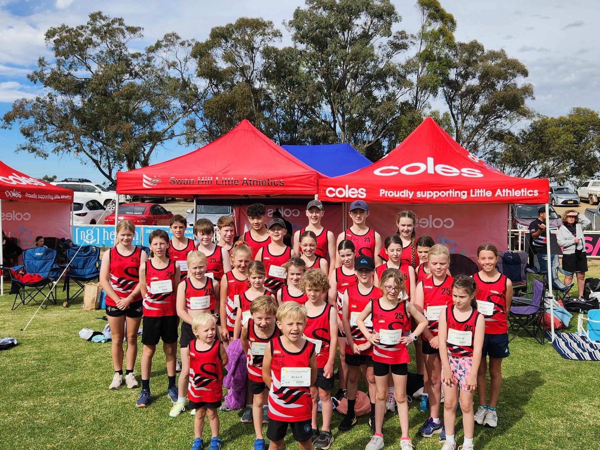 A youth running team in red uniforms poses under sponsor tents on a grassy field.