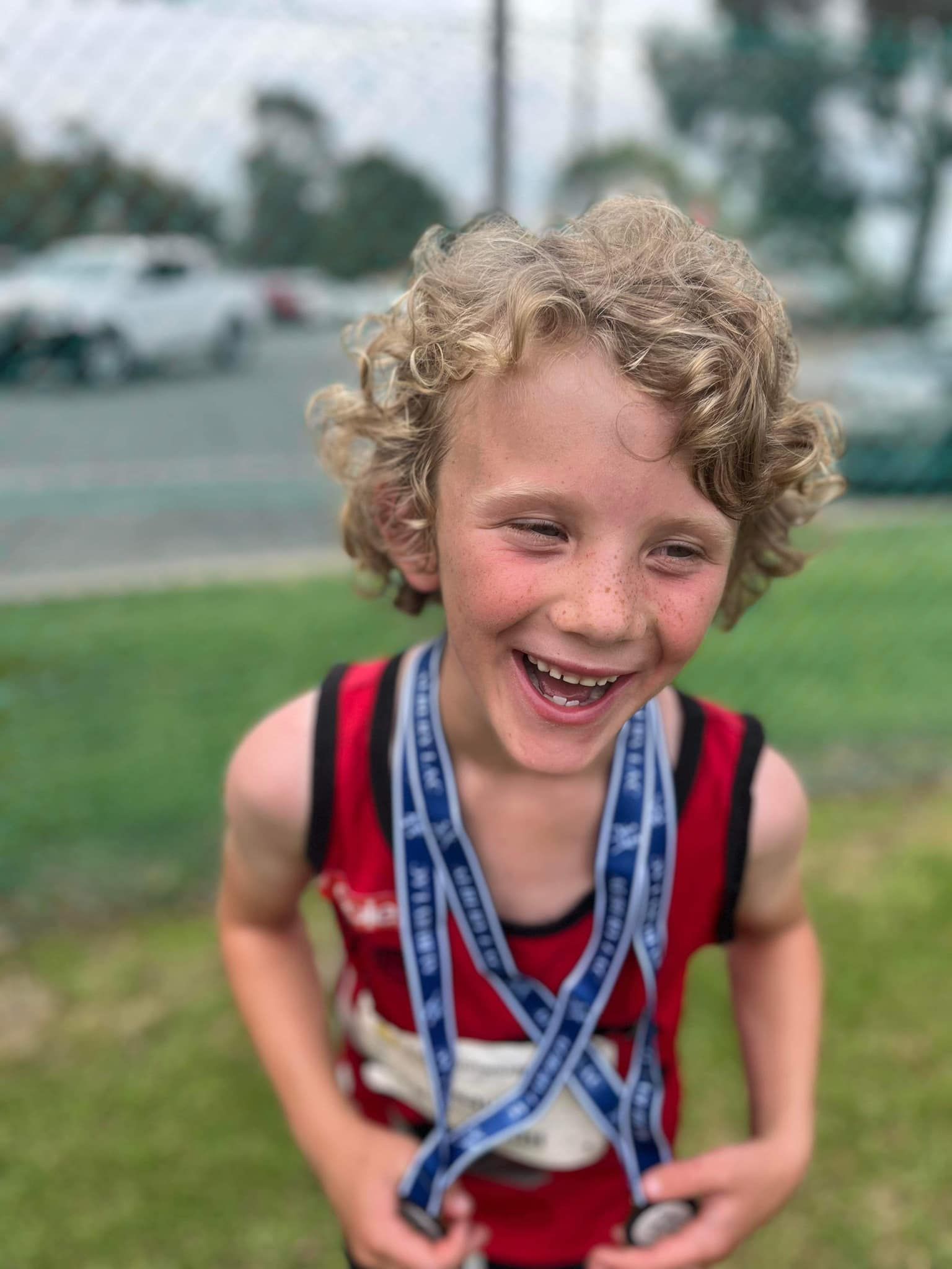 Boy with curly blonde hair smiles, wearing medals around his neck, red and white singlet on a grassy field.