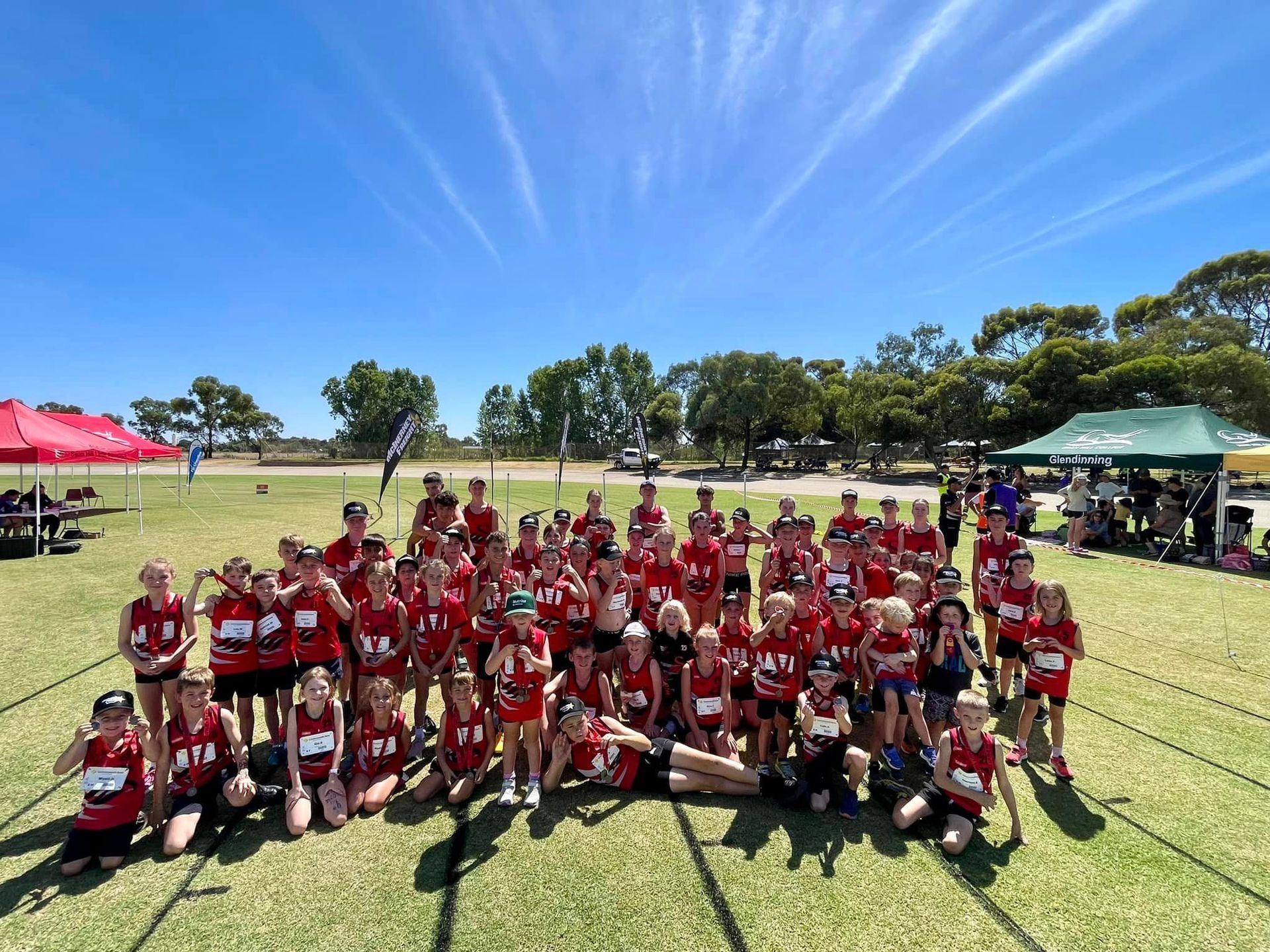 Large group of runners in red uniforms pose on a grassy field under a blue sky.