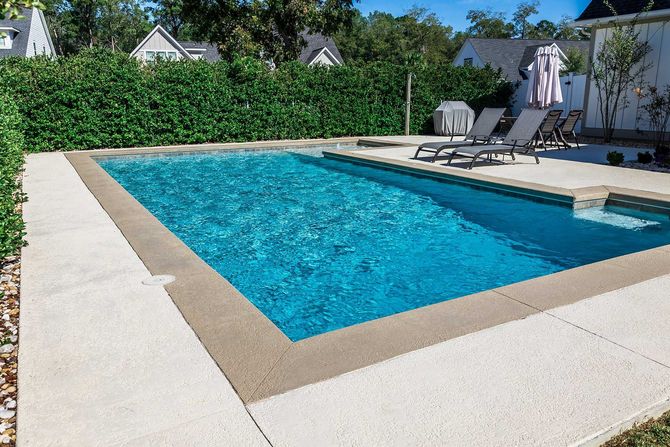 A rectangular new swimming pool with tan concrete edges in the fenced backyard of a new construction house 
