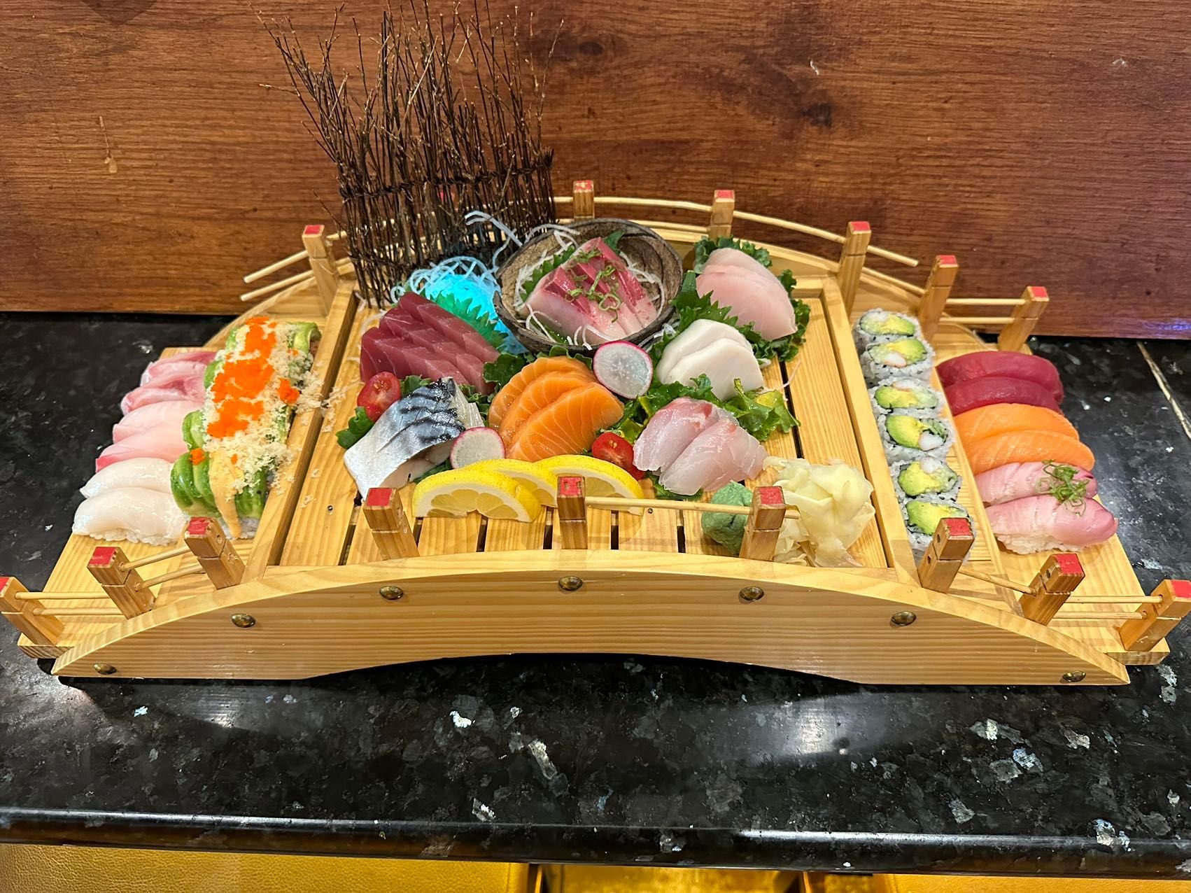 Sushi platter arranged on a wooden bridge-shaped tray, with various sashimi, rolls, and garnishes.