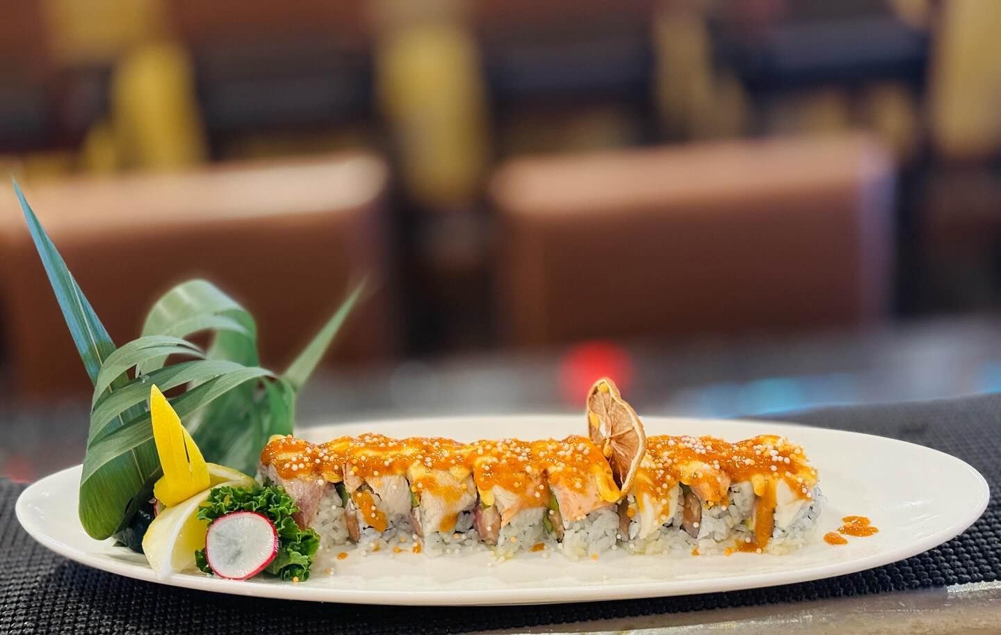 Sushi roll with orange sauce on a white plate, garnished with green leaves and radish, restaurant setting.