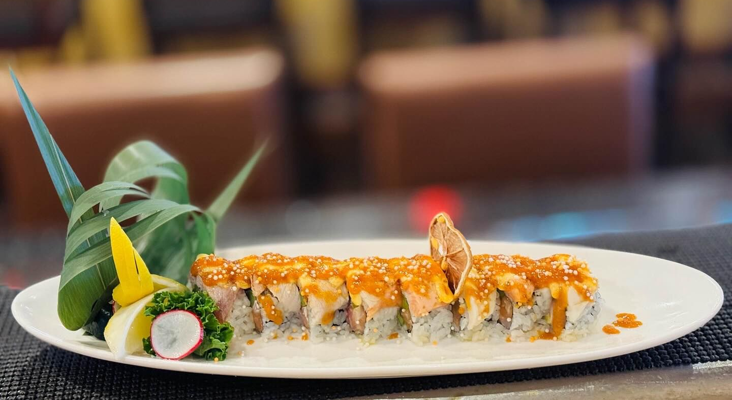 Sushi roll on a white plate, garnished with greens and sauce, in a restaurant setting.