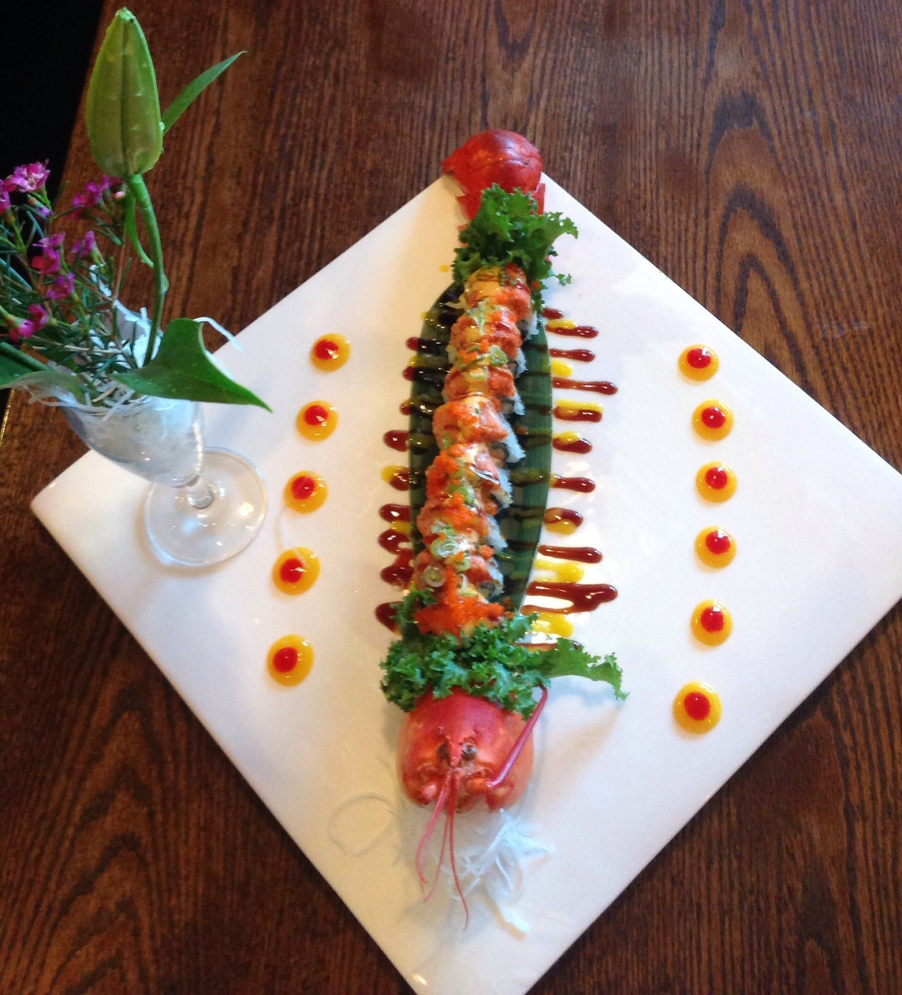 Sushi roll artfully arranged on a square white plate, with decorative sauce dots and flower garnish.