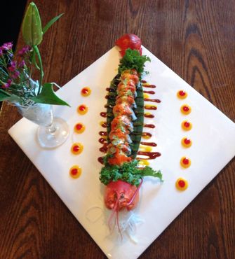 Sushi roll artfully arranged on a white plate, garnished with vegetables and sauces, alongside a floral arrangement.