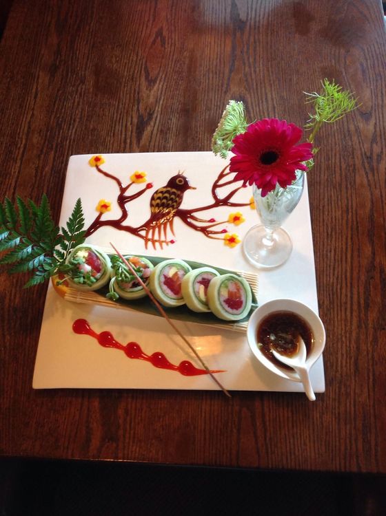 Sushi roll arrangement on a white plate with a bird design, flowers, and dipping sauce.