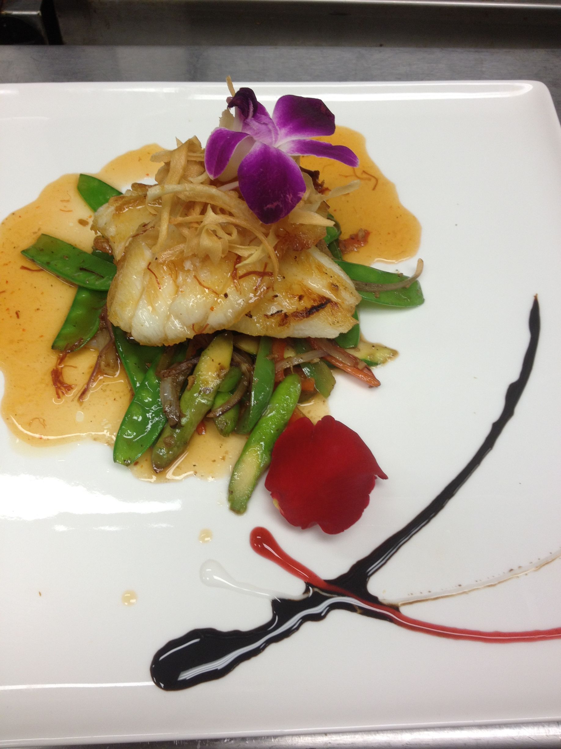 Gourmet dish: white fish atop asparagus, snap peas, sauce, and onion crisps, with orchid flower and artistic sauce swirls on white plate.