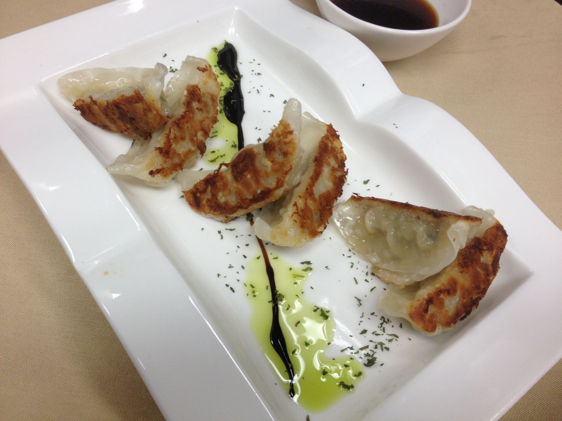Pan-fried dumplings on a white plate with a dark sauce. Green garnish and small bowl of sauce.