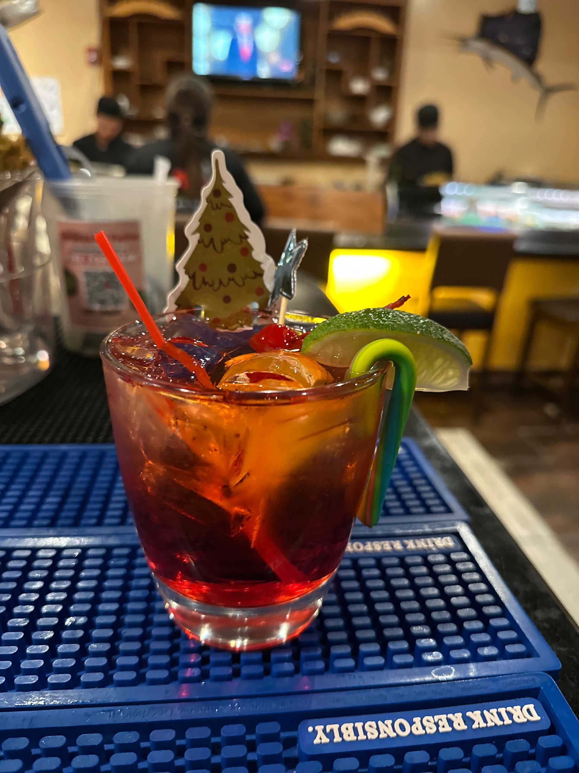 Cocktail with red and amber layers, garnished with a lime, cherry, and Christmas tree decoration, set on a blue bar mat.
