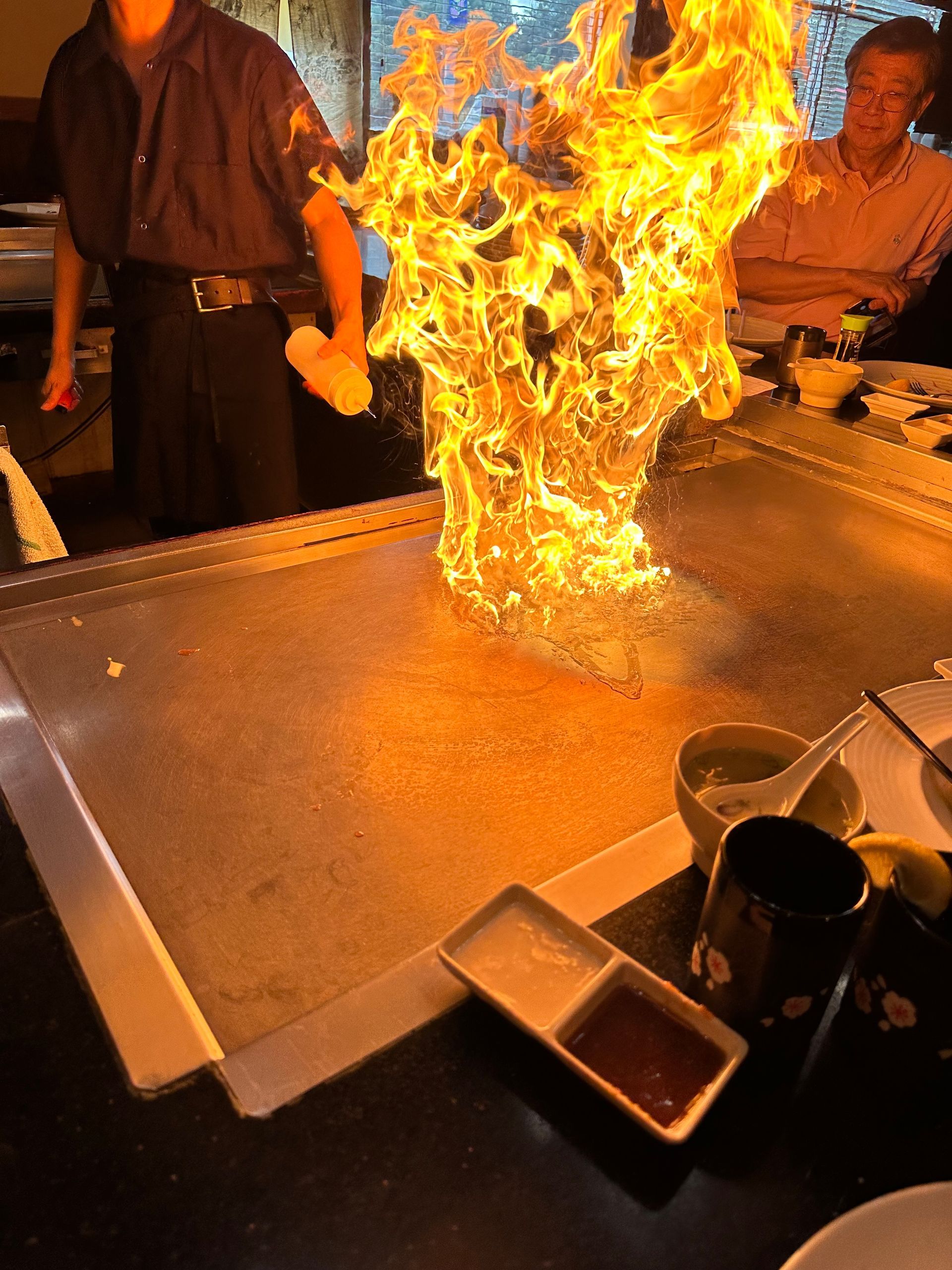 Chef ignites a large flame on a teppanyaki grill. Restaurant setting, people watch.