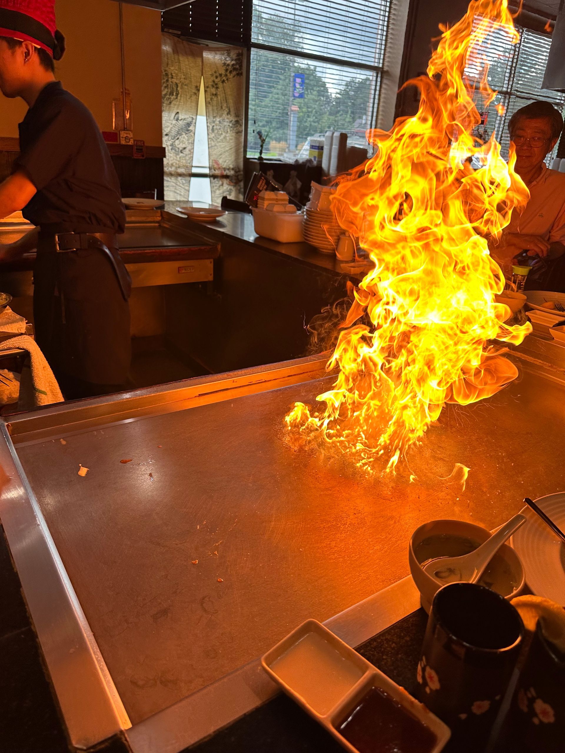 Teppanyaki chef ignites a fire on the grill at a restaurant, with diners in the background.