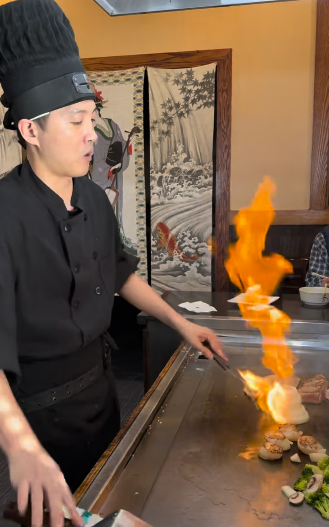 Chef at a hibachi grill, igniting flames over food on a hot surface.