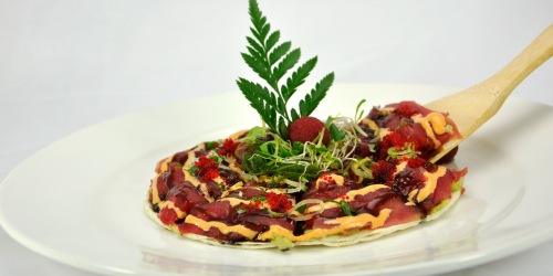 Sushi on a white plate, topped with sauce, microgreens and fern. A wooden spoon holds a portion.