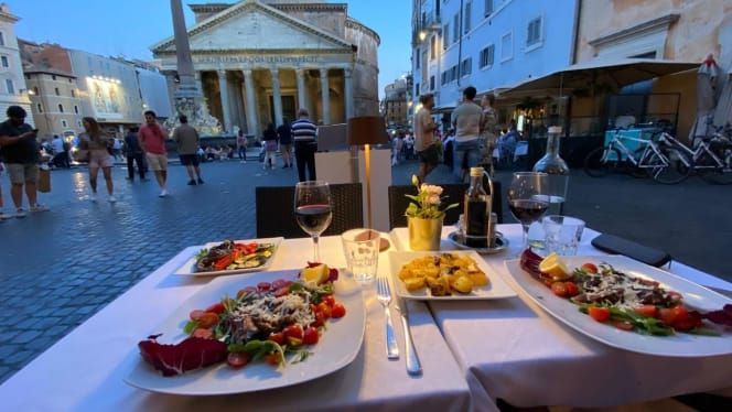 Tavolo apparecchiato per la cena con il Pantheon di Roma, Italia, sullo sfondo; piatti, bicchieri da vino e persone.