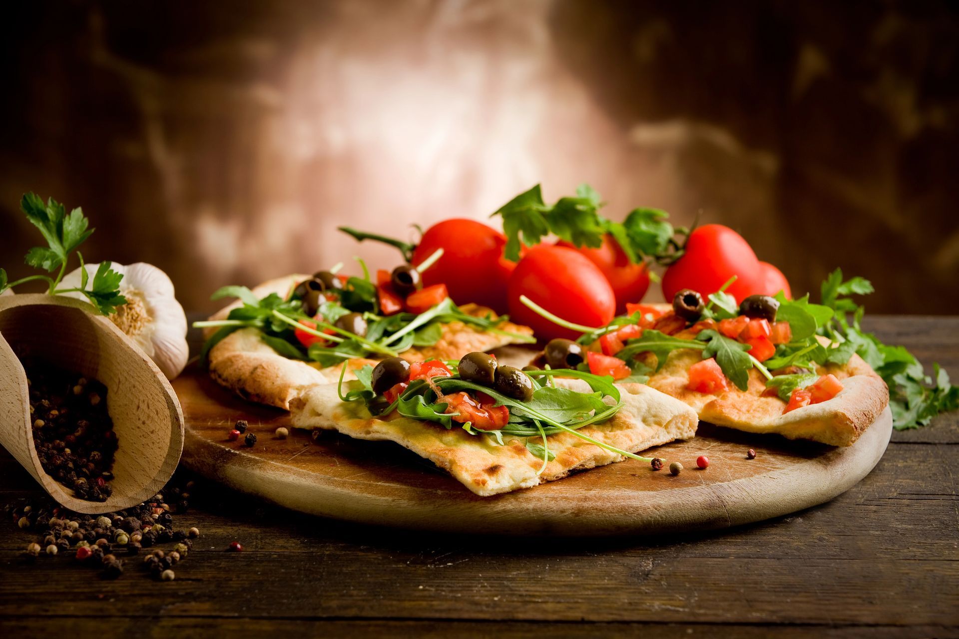 Pizza condita con pomodori, olive e rucola su un tagliere di legno. Aglio e spezie a portata di mano.