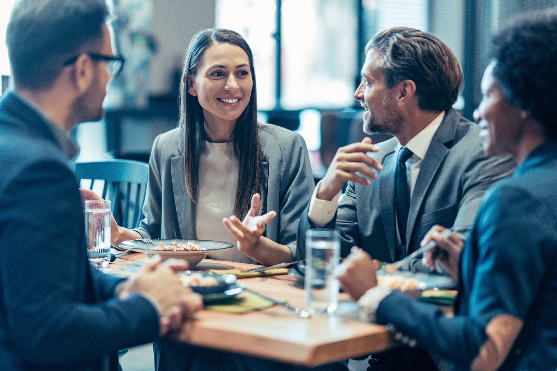 Quattro professionisti in giacca e cravatta in un ristorante, conversano e sorridono seduti a un tavolo con del cibo.