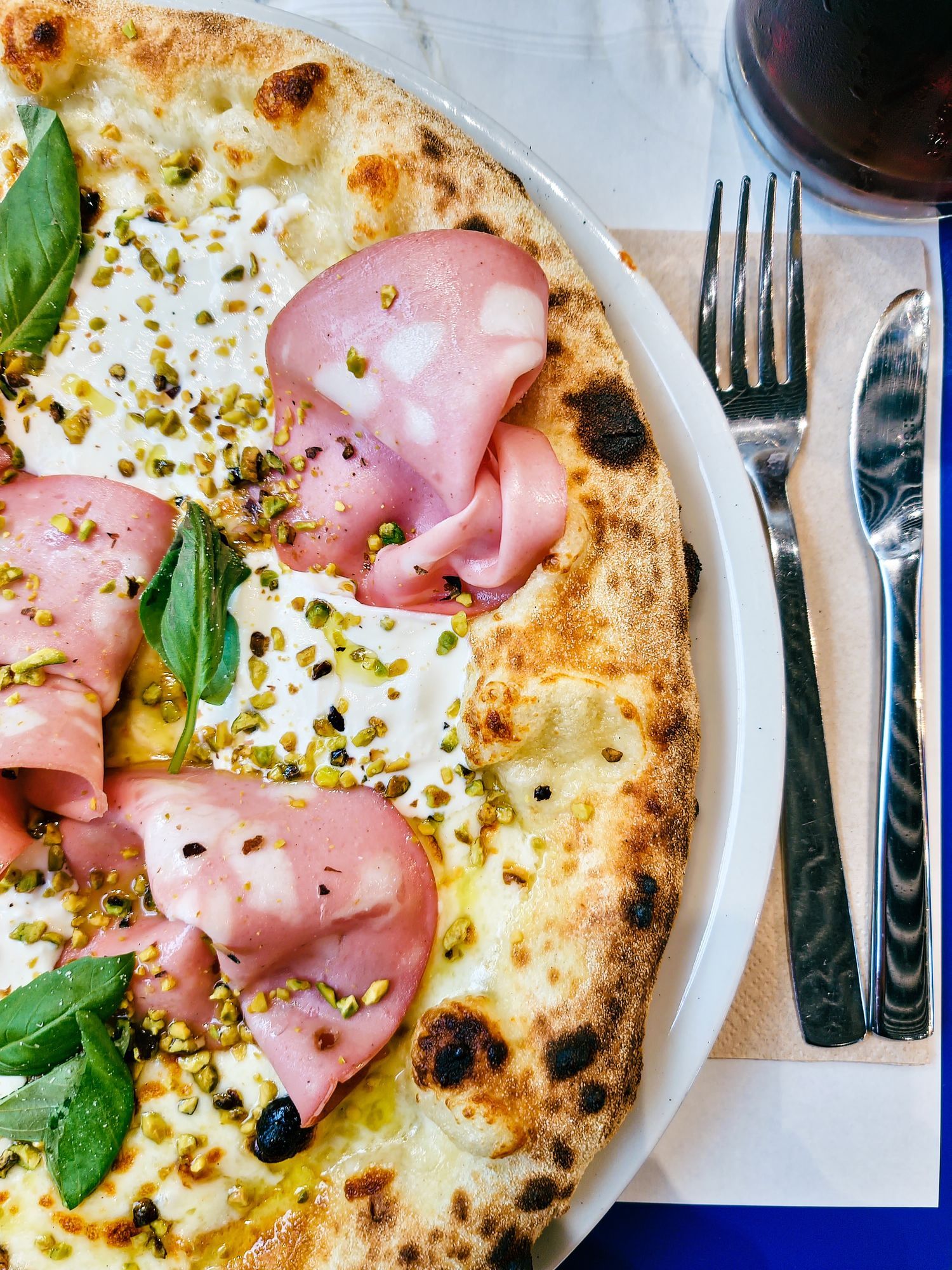 Pizza con mortadella, pistacchi e basilico. Servita su un piatto bianco con posate.