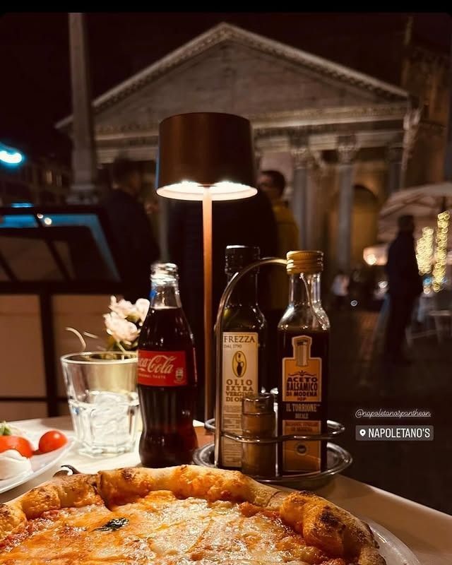 Pizza, Coca-Cola e bottiglie di olio sul tavolo di un ristorante di notte, con un edificio storico sullo sfondo.