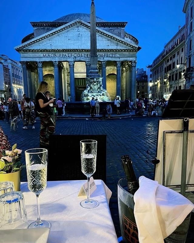 Bicchieri di champagne su un tavolo, sullo sfondo il Pantheon di Roma al tramonto. La gente si raduna in piazza.