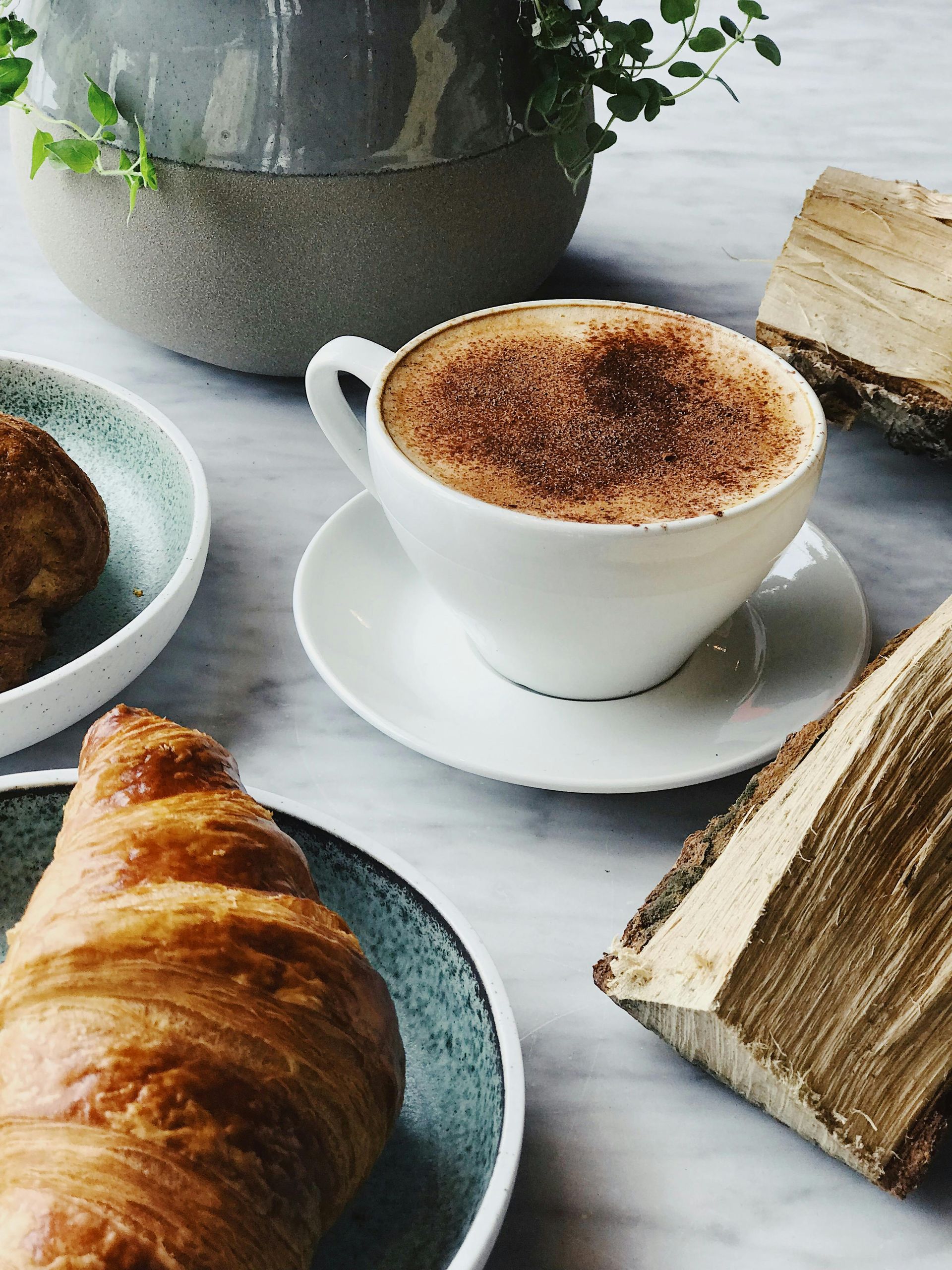 Cappuccino in tazza bianca su piattino con croissant, pasticcini su piatti blu e pezzi di legno.