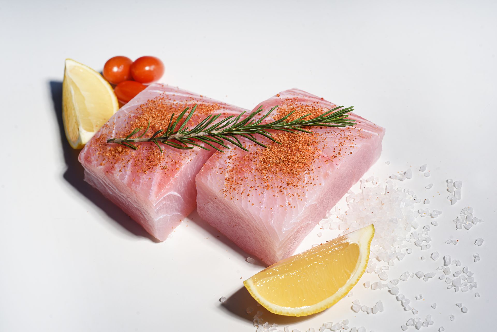Two raw fish fillets seasoned with herbs and spices, garnished with lemon wedges, cherry tomatoes, and scattered salt.