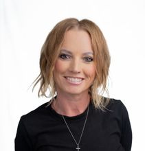A smiling person with shoulder-length blonde hair, wearing a black shirt and a silver cross necklace against a white backdrop.