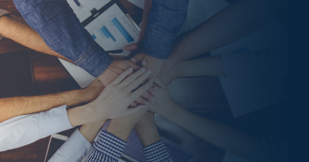 A top-down view of several people stacking their hands together in a gesture of teamwork over a desk with documents.