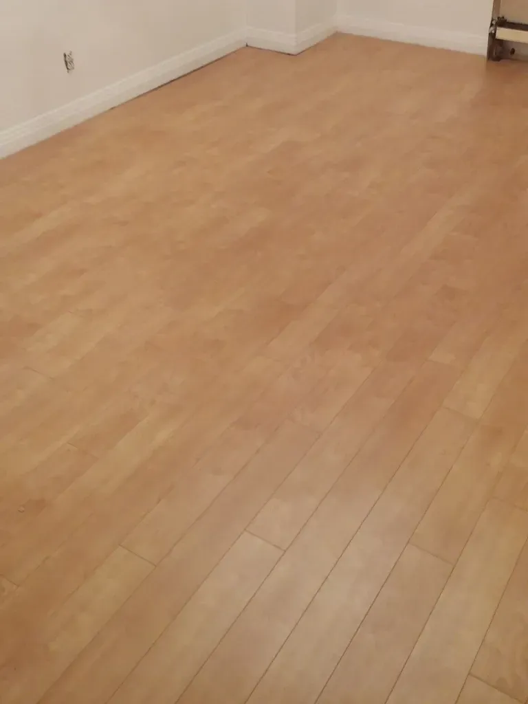 Revêtement de sol stratifié clair à l'aspect bois dans une pièce aux plinthes et murs blancs.