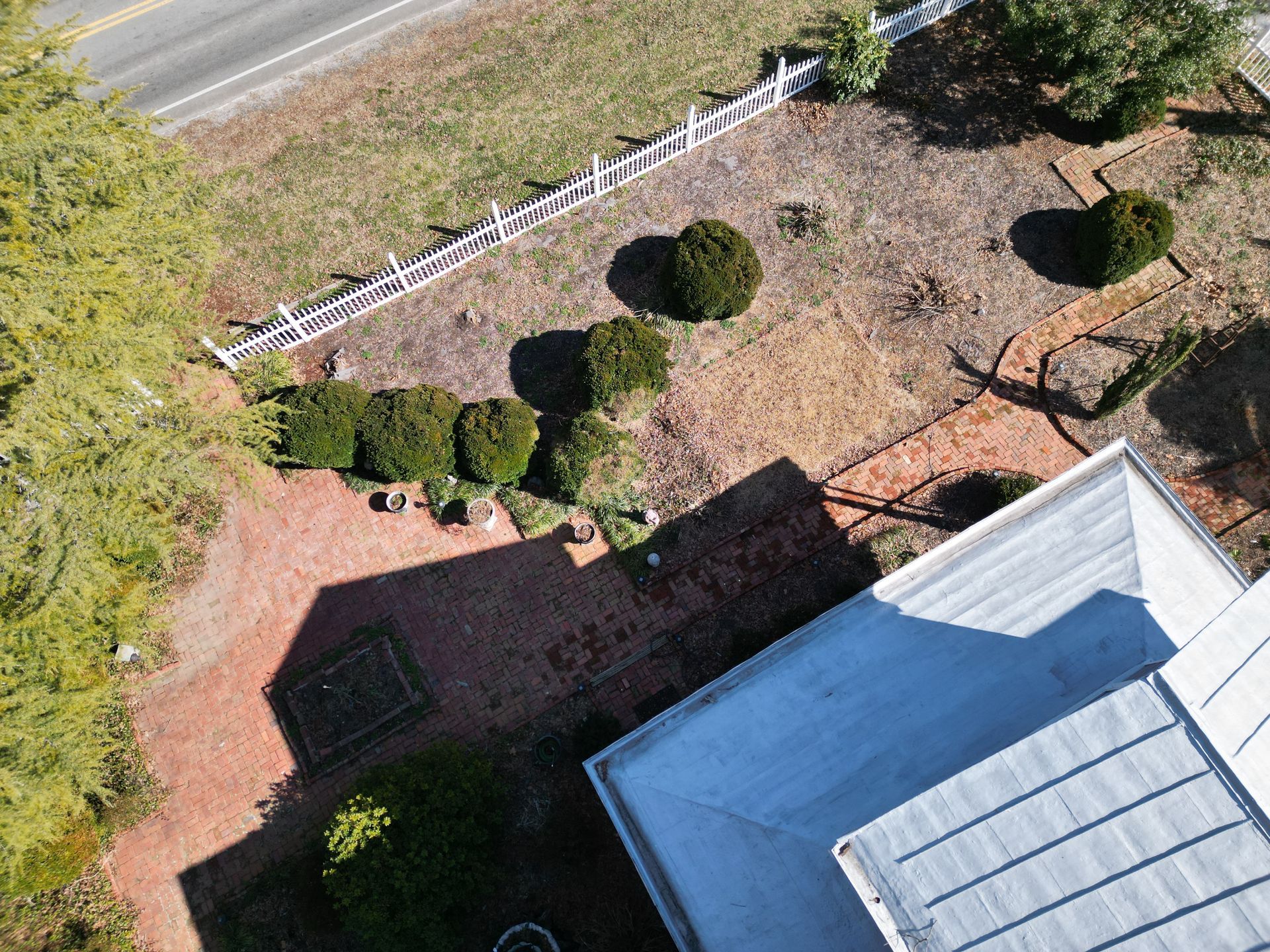 Aerial view of a house and yard with bushes and white fence, beside a road.