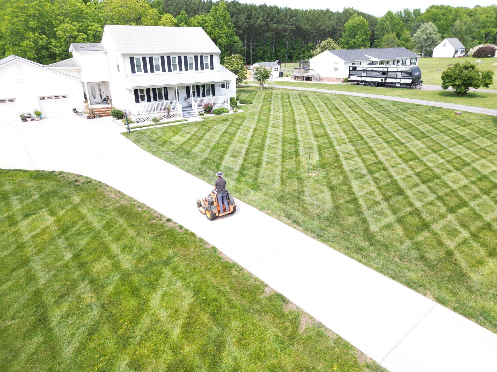 Person mowing lawn on a riding mower, driveway leading to white house, grid-patterned lawn, sunny day.
