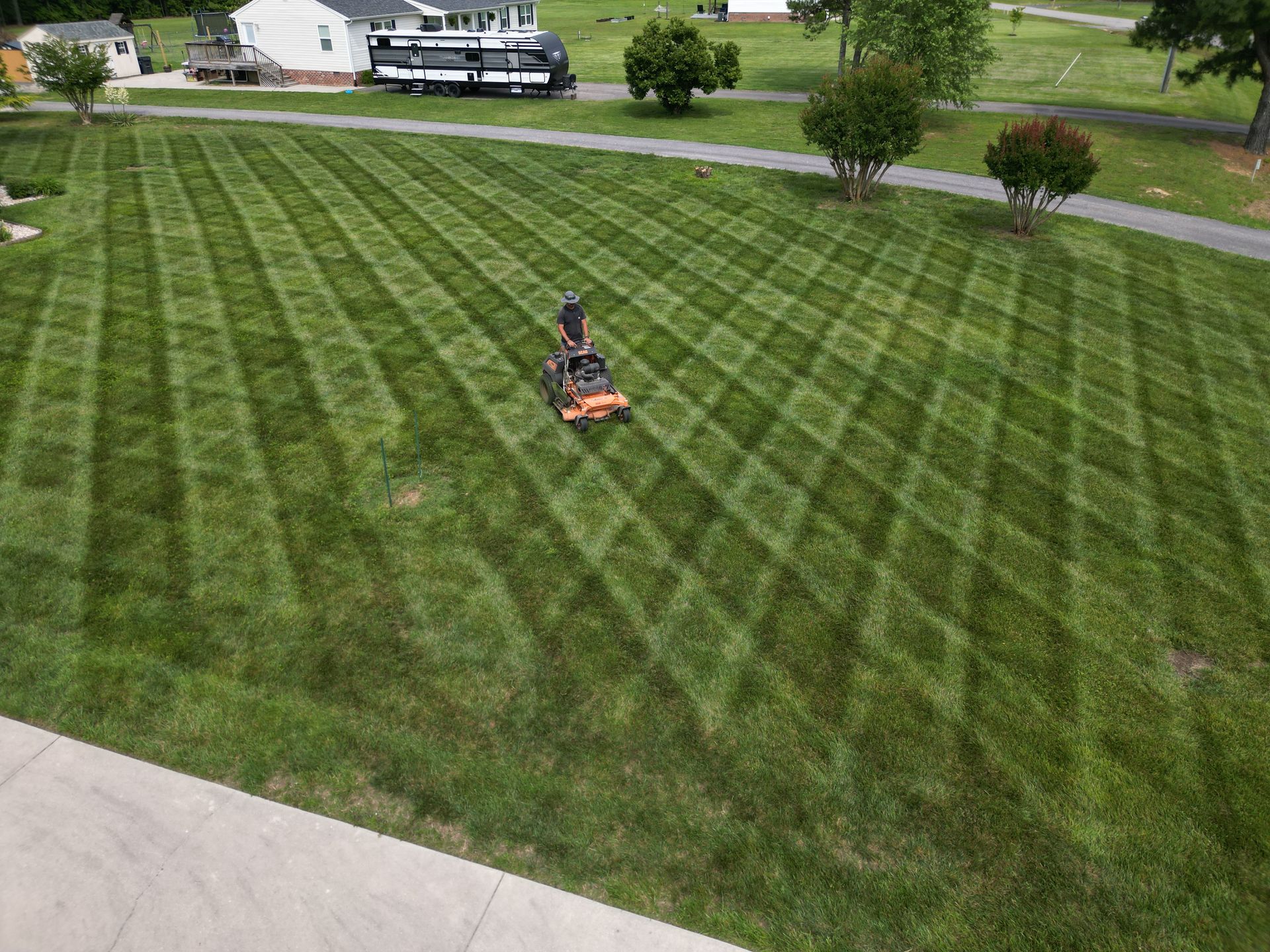 Lawnmower cutting a diamond pattern into a green lawn, with a house and trees in the background.